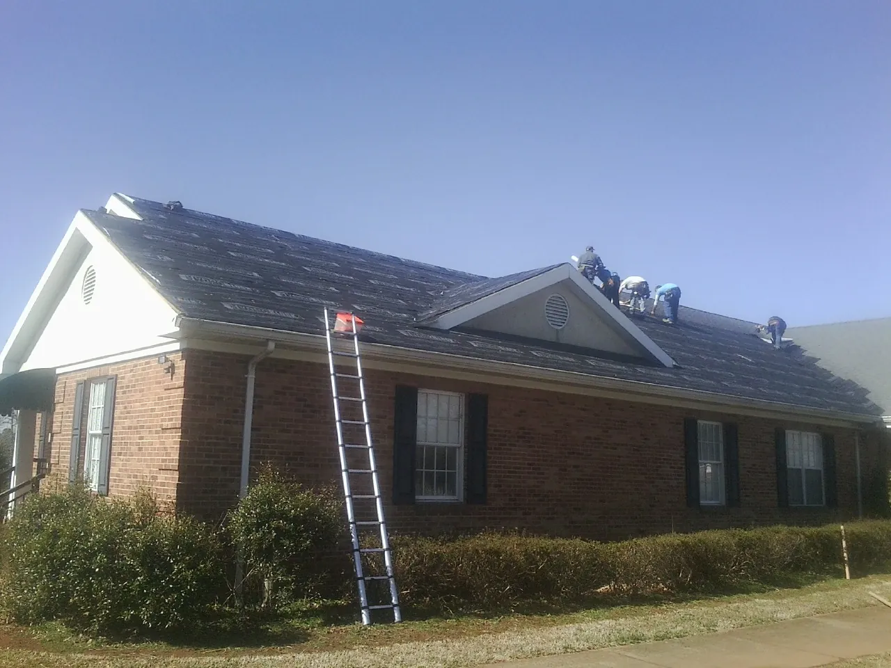 Crew tearing shingles off a brick ranch roof with a ladder set — Metal Roof Repair work by our crew in Scotland Neck