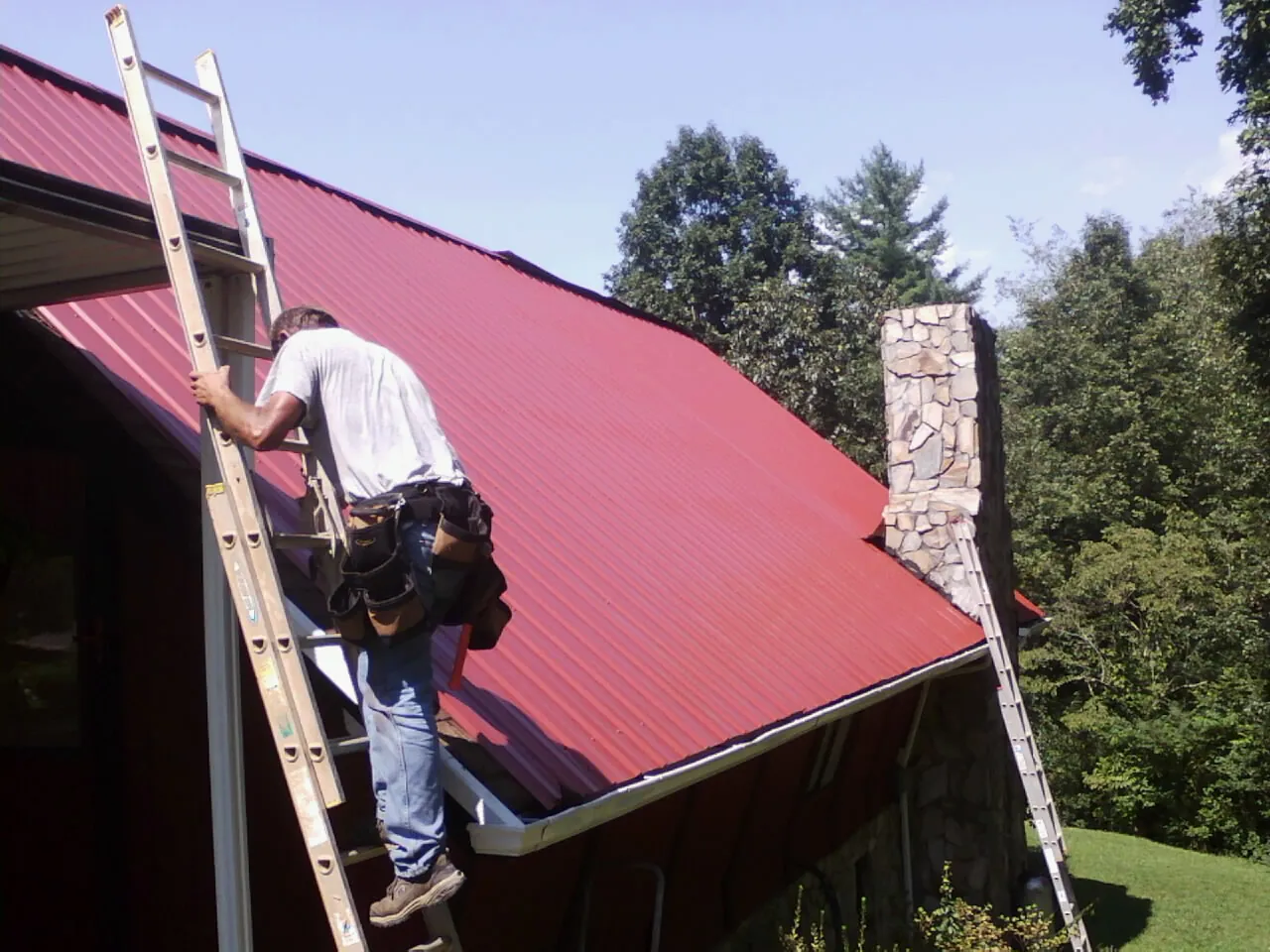 Roofer on a ladder beside a red metal roof and stone chimney — Soffit & Fascia Repair by Roofing Brothers in Scotland Neck